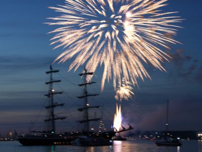 Am dunkelblauen Nachthimmel explodiert ein Feuerwerkskörper. Im Vordergrund auf dem Wasser sieht man die Silhoutte eines 2-Mast-Segelschiffes.