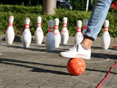 Weiße Bowlingkugeln stehen aufgereiht auf Asphalt. Davor liegt eine orangene Bowlingkugel, die von einem Fuß in weißem Turnschuh in Position gehalten wird.