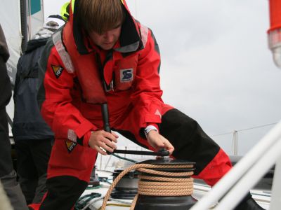 Ein Segler  in rotem Segelanzug legt Segeltau um eine Winsch und kurbelt.