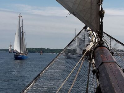 Blick über einen Klüverbaum mit Klüvernetz. Im Hintergrund ein Segelschiff mit weißen Segeln auf dem Wasser.