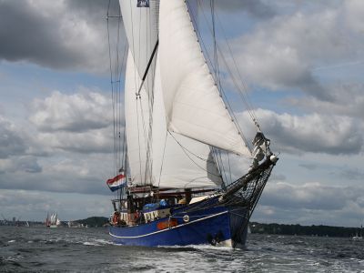 Ein Segelschiff mit blauem Rumpf und vollen weißen Segeln segelt auf dem Wasser. Am Himmel sind weiß-graue Wolken.