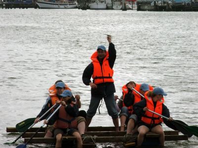 Ein Mann in roter Schwimmweste steht auf einem schwimmenden Floß und reckt die Faust siegreich in die Luft