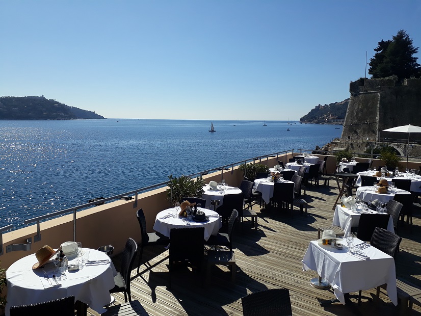 Blick auf eine Terrasse direkt am Meer, auf der Tische mit weißen Tischdecken stehen. Im Hintergrund sieht man blauen Himmel und blaues Meer in einer von zwei Felsen eingeschlossenen Bucht.