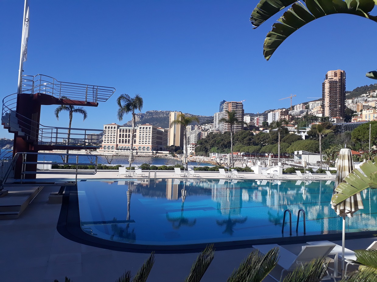 Im Vordergrund ist ein Pool mit einem Dreimeterturm. Im Wasser des Schwimmbeckens spiegelt sich die Bucht von Monaco.