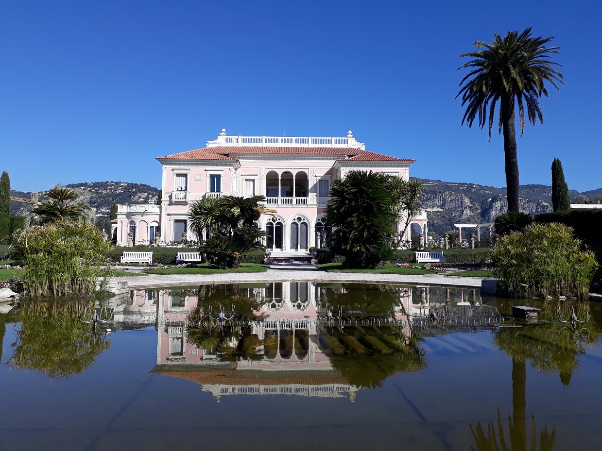 Die Villa Rothschild im Renaissance-Stil spiegelt sich in einem See, der vor der Villa liegt. Am rechten Seeufer steht eine hohe Palme.